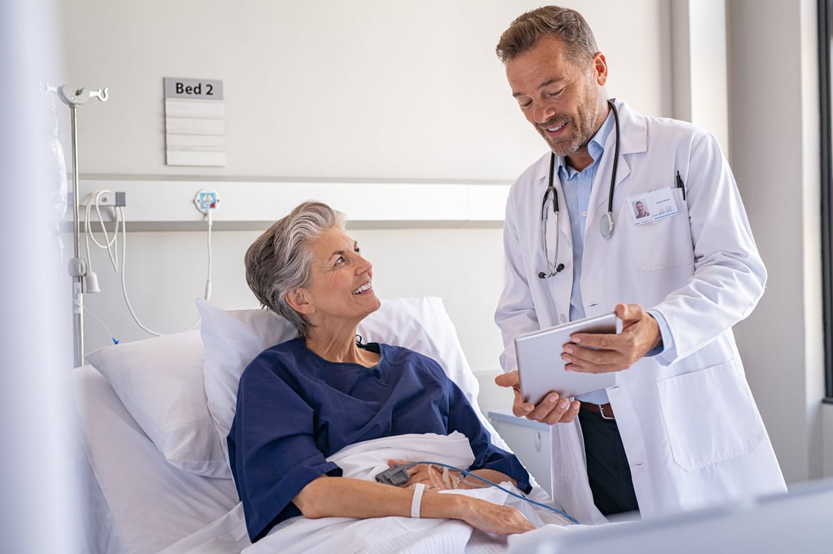 A smiling senior patient on a hospital bed chatting with a confident surgeon who holds a digital tablet